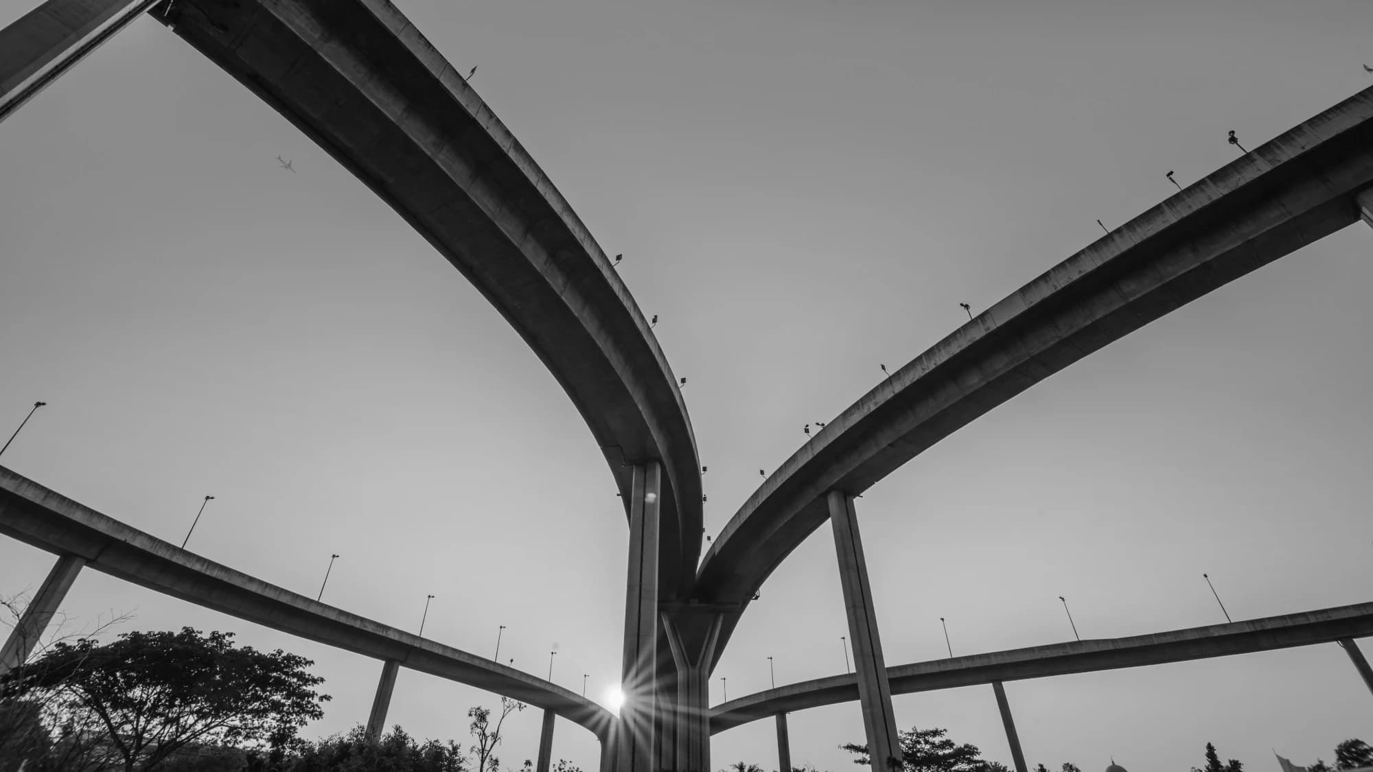 Curving elevated highway interchange ramps converging against a pale sky, sun flaring below, monochrome