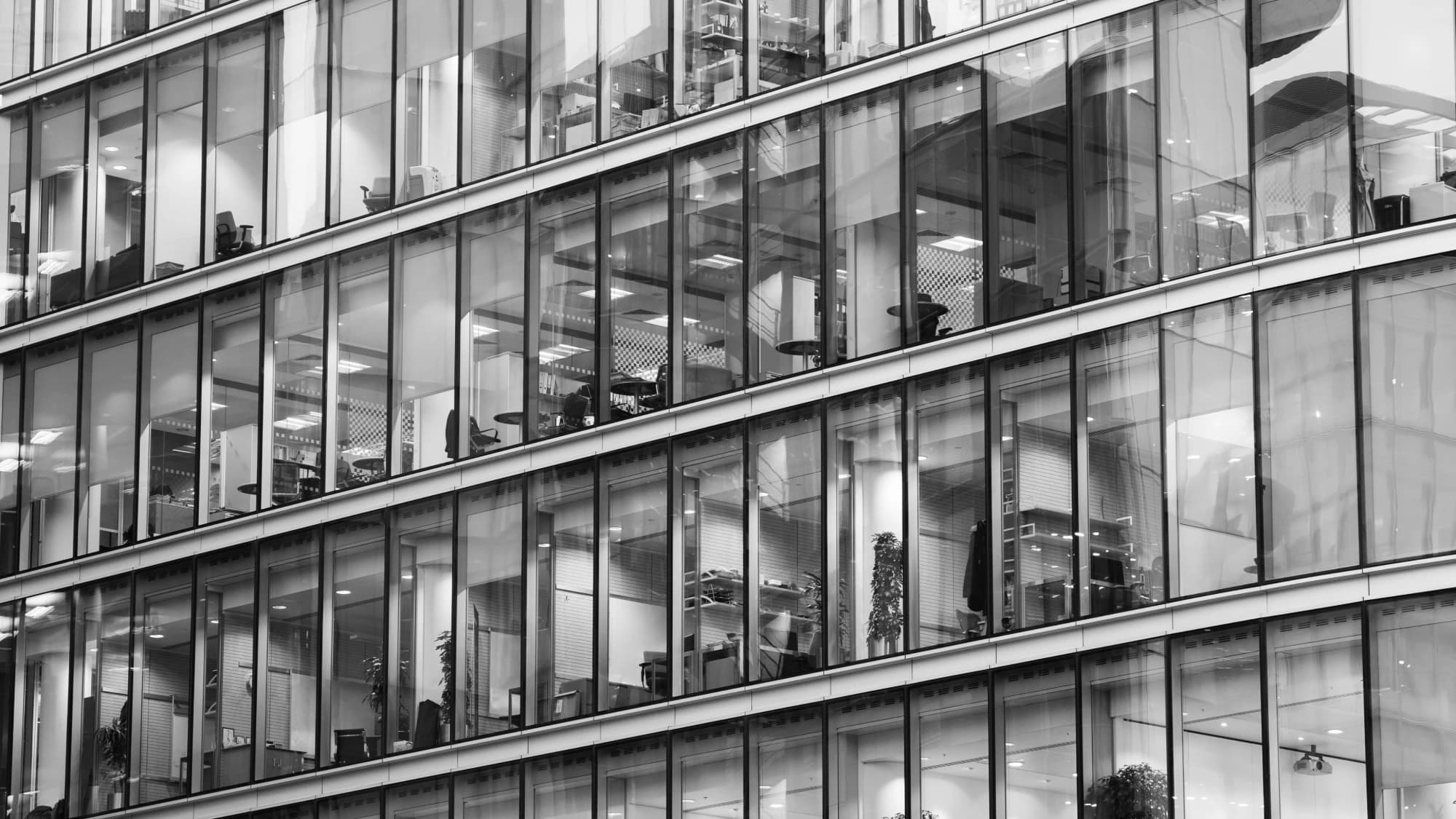 Glass facade of an office building with lit interior floors and workspaces, monochrome