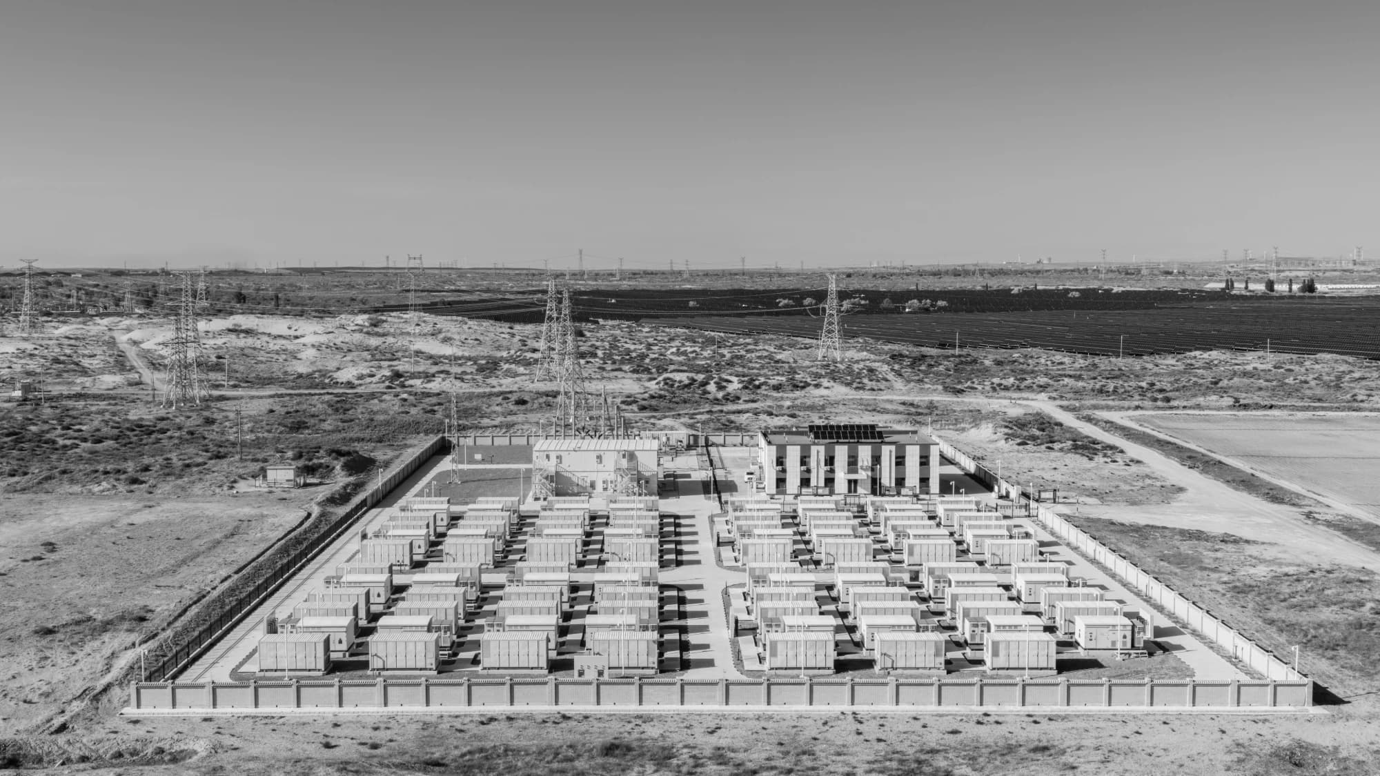 Aerial view of a grid-scale battery storage yard beside transmission towers and a solar field, monochrome