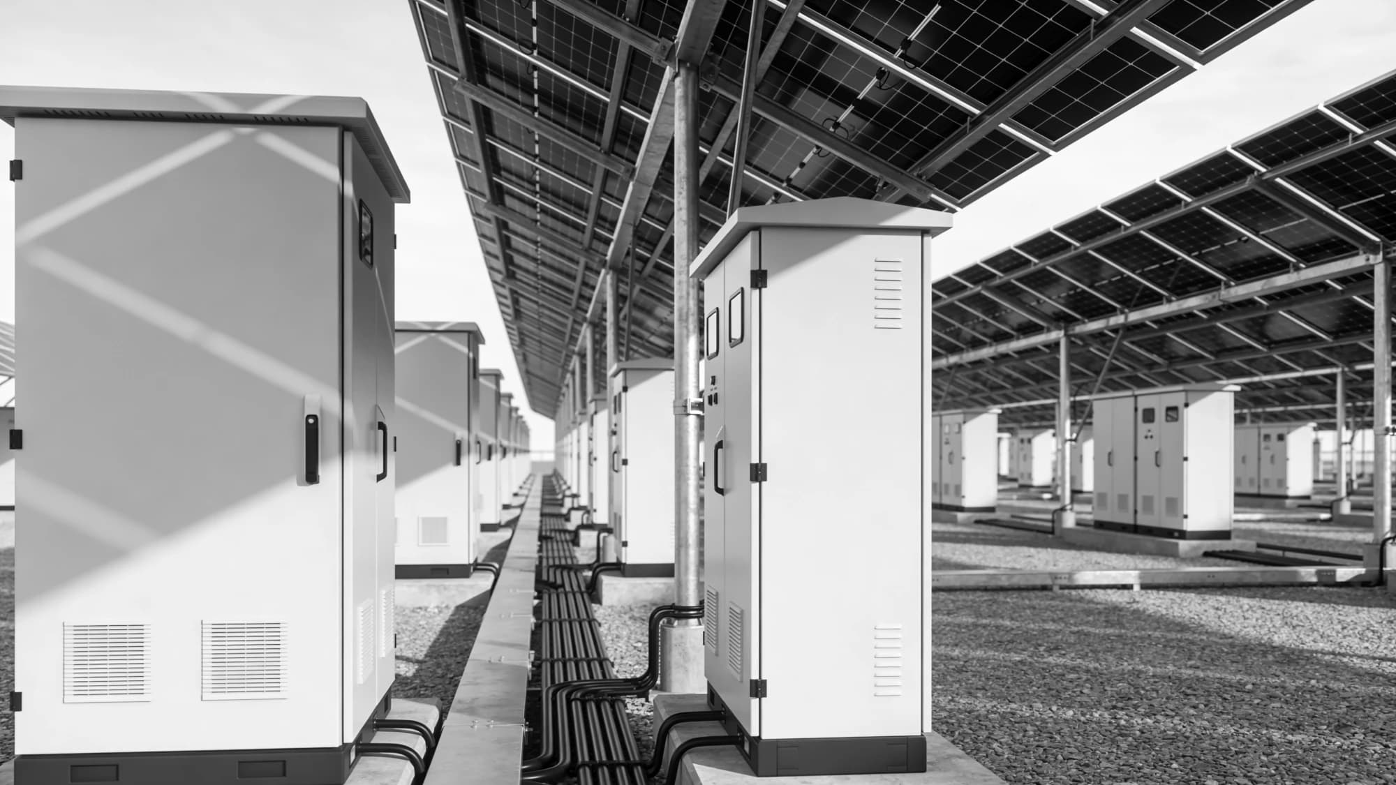 Rows of battery storage and inverter cabinets beneath a solar panel canopy, receding to the horizon, monochrome